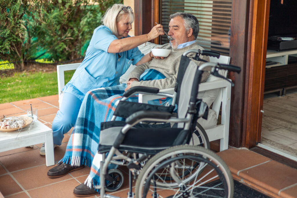 Nurse assisting elderly man eating in wheelchair on patio